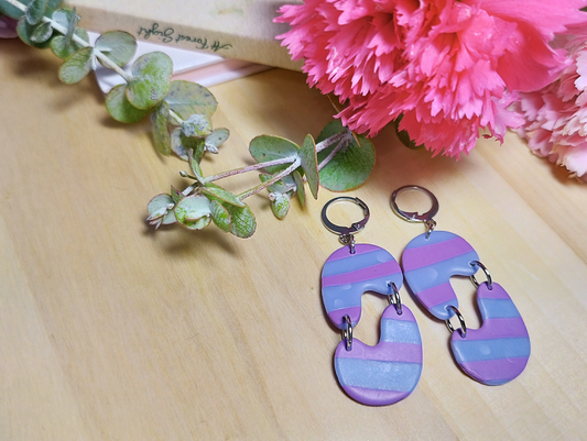 Rayado Huggies, Handmade Polymer Clay Earrings. The base is a Silver colour Huggies attached with a silver colour jump ring to an half abstract Arch shape that is connected with jump rings with another half abstract Arch shape both in the colour of Periwinkle and Deep Lilac.